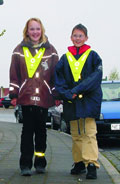 Zwei Kinder stehen nebeneinander auf einem Bürgersteig. Beide tragen leuchtende Warnwesten, die über ihrer Alltagskleidung angezogen sind, um die Sichtbarkeit im Straßenverkehr zu erhöhen. Im Hintergrund sind Autos geparkt. Das Bild betont Arbeitsschutz und Prävention für die Unfallkasse NRW, indem es die Wichtigkeit von Reflexkleidung im Straßenverkehr demonstriert.