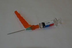 Eine leere Spritze mit einem orangen Sicherheitsmechanismus, der den Nadelbereich abdeckt, liegt auf einem neutralen Hintergrund. Der Mechanismus ist halb geöffnet und zeigt, wie er die Nadel schützt. Diese Art von Spritzen wird verwendet, um Verletzungen im medizinischen und pflegerischen Bereich zu vermeiden und die Sicherheit der Mitarbeiter zu gewährleisten.