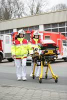 Zwei in Schutzkleidung gekleidete Sanitäter schieben eine Trage vor einem großen roten Rettungswagen. Sie befinden sich auf einem gepflasterten Parkplatz vor einem Gebäude mit grauem Dach. Bäume sind im Hintergrund sichtbar. Diese Szene steht im Kontext des Arbeitsschutzes und der Unfallprävention durch die Unfallkasse NRW.