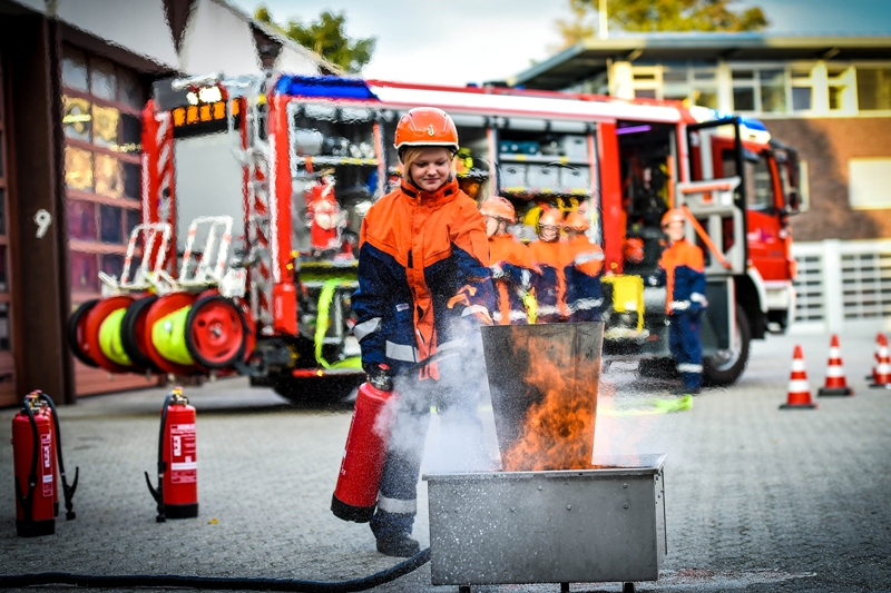 Eine Gruppe von Kindern, die orange Schutzkleidung und Helme trägt, führt eine Feuerlöschübung vor. Eines der Kinder hält einen Feuerlöscher auf ein brennendes Objekt, das auf einem gepflasterten Hof steht. Im Hintergrund befindet sich ein Feuerwehrauto mit Feuerwehrzubehör und mehrere Feuerlöscher sind in der Nähe platziert. Die Szene vermittelt die Bedeutung von Präventionsmaßnahmen und Arbeitsschutz im Kontext der Unfallkasse NRW.
