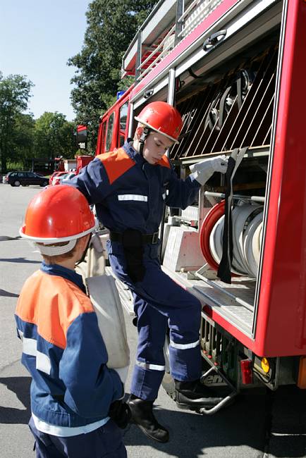 Zwei Feuerwehrleute mit orangenen Helmen und blauer Schutzkleidung stehen an einem offenen Einsatzfahrzeug. Eine Person entnimmt Schläuche aus dem Fahrzeug, während die andere zuschaut. Im Hintergrund sind Bäume und weitere Fahrzeuge zu erkennen. Diese Szene zeigt wichtige Aspekte der Arbeitssicherheit und Prävention im Einsatzalltag der Feuerwehr.