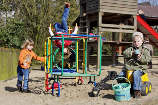 Drei Kinder spielen auf einem Spielplatz in einem großen Sandkasten. Links steht ein Kind mit einem orangefarbenen Pullover neben einem bunten Klettergerüst. In der Mitte hängt ein Kind kopfüber an dem Klettergerüst und blickt lächelnd zu den anderen Kindern. Rechts sitzt ein Kind mit einer Mütze auf einem gelben Spielzeug-Bagger und hält eine grüne Plastikschaufel und einen Eimer. Im Hintergrund befindet sich ein Holzturm mit einer roten Rutsche. Der Kontext passt zur Unfallkasse NRW im Bereich Arbeitsschutz und Prävention, da das korrekte und sichere Spielverhalten im Fokus steht.
