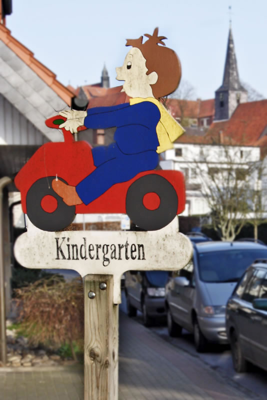 Ein Holzschild zeigt ein stilisiertes Kind mit braunem Haar auf einem roten Spielzeugauto. Das Kind trägt blau-gelbe Kleidung. Darunter steht in Großbuchstaben 'Kindergarten'. Im Hintergrund sind geparkte Autos, ein Gebäude mit roten Ziegeln und der Kirchturm einer Stadt zu sehen. Das Schild dient zur Verkehrswarnung und Erhöhung der Sicherheit in der Nähe eines Kindergartens, passend zum Kontext der Unfallkasse NRW für Arbeitsschutz und Prävention.