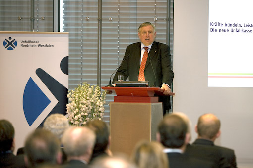 Ein Mann steht am Rednerpult und hält eine Rede bei einer Veranstaltung der Unfallkasse Nordrhein-Westfalen. Im Hintergrund sind ein Banner mit dem Logo der Unfallkasse NRW und ein Bildschirm mit Präsentationstext zu sehen. Vor dem Redner sitzen mehrere Zuhörer. Der Fokus liegt auf Themen wie Arbeitsschutz und Prävention.