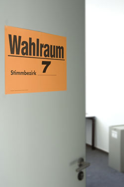 Eine leicht geöffnete graue Tür zeigt ein orangefarbenes Schild mit der Aufschrift 'Wahlraum 7 – Stimmbezirk'. Der Raum im Hintergrund wirkt unbesetzt und beinhaltet einen großen, grauen Behälter. Die Szene vermittelt Vorsichtsmaßnahmen und Orientierung bei der Organisation von Wahleinrichtungen, relevant für Arbeitsschutz und Prävention.