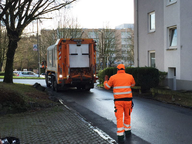 Ein Müllarbeiter in orangefarbener Schutzkleidung mit Reflexstreifen dirigiert ein Müllfahrzeug sicher rückwärts auf einer schmalen Straße. Der Arbeiter hebt beide Arme, um dem Fahrer Anweisungen zu geben. Im Hintergrund sind Bäume und ein mehrstöckiges Gebäude zu sehen. Die Szene betont Arbeitsschutz und Prävention im öffentlichen Raum.