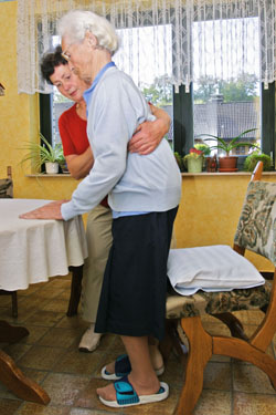 Eine Pflegekraft in rotem Oberteil unterstützt eine ältere Frau mit weißem Haar, die beim Aufstehen von einem Stuhl Hilfe benötigt. Der Raum hat gelbe Wände und es stehen Pflanzen auf der Fensterbank. Die Szene zeigt den Kontext der Unfallkasse NRW in Bezug auf Arbeitsschutz und Prävention in der Pflege.