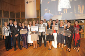 Eine große Gruppe von Menschen steht auf einer Bühne bei einer Preisverleihung, die möglicherweise von der Unfallkasse NRW für besondere Verdienste im Bereich Arbeitsschutz und Prävention veranstaltet wird. Die Personen halten Schilder oder Auszeichnungen in den Händen. Im Hintergrund sind Bühnenlichter und ein Banner mit dem Logo der Organisation zu erkennen.