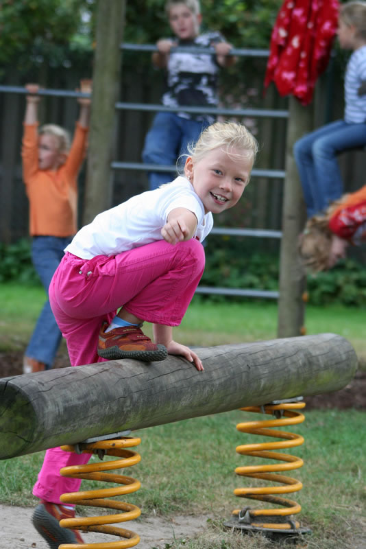 Ein blondes Mädchen in rosa Hose und weißem T-Shirt balanciert auf einer Federwippe aus Holz auf einem Spielplatz. Die Federwippe ist gelb und hat zwei Federn. Im Hintergrund klettern Kinder auf einem Klettergerüst. Der Spielplatz ist von einem Holzzaun umgeben und von grüner Vegetation gesäumt. Das Bild steht im Kontext der Unfallkasse NRW, die Arbeitsschutz und Prävention fördert.