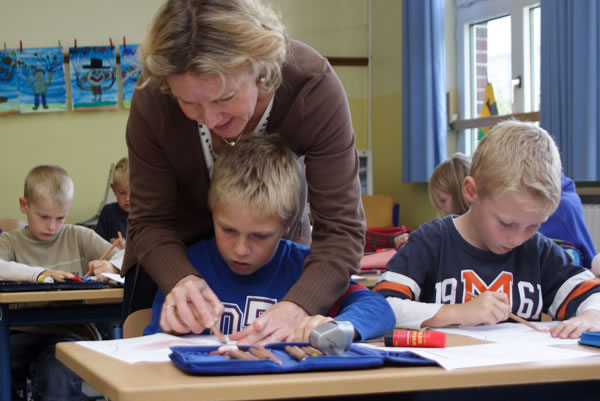 Eine Lehrerin beugt sich über einen Jungen, um ihm und einem anderen Schüler bei einer Aufgabe zu helfen. Die beiden Jungen sitzen an einem Tisch mit Malutensilien und konzentrieren sich auf ihre Arbeit. Im Hintergrund sind weitere Kinder zu sehen, die ebenfalls an ihren Tischen arbeiten. Das Klassenzimmer hat große Fenster, durch die Tageslicht einfällt. Hinweis auf Arbeitsschutz und Prävention: Die Lehrerin sorgt für individuelle Unterstützung und fördert ein sicheres Lernumfeld.