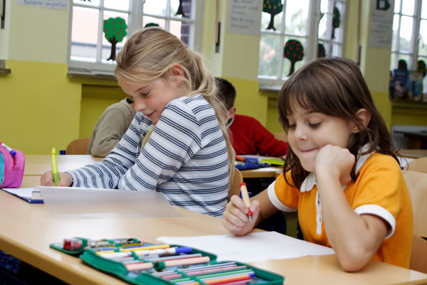 Zwei junge Mädchen sitzen an Schreibtischen in einem Klassenzimmer und schreiben auf Papier. Im Vordergrund liegt eine Federtasche mit Buntstiften. Die gelben Wände sind mit kindlichen Arbeiten und Informationen dekoriert. Das Bild betont die Bedeutung von Sicherheit und Komfort bei der Arbeit von Kindern in Bildungsumgebungen.
