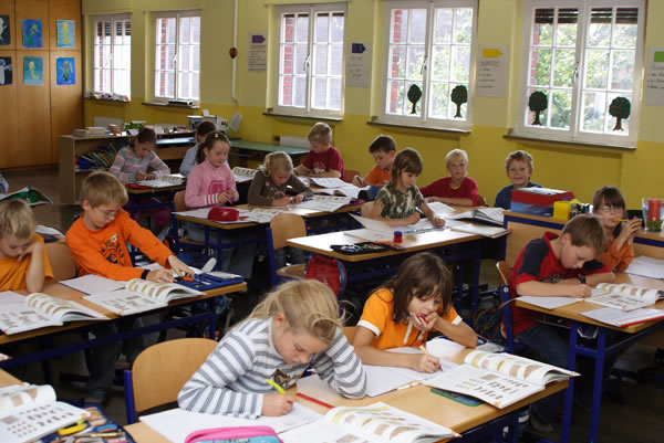 Eine Gruppe von Kindern sitzt in einem hellen Klassenraum an einzelnen Tischen. Sie sind in Aktivitäten vertieft, wie das Schreiben oder Lesen in Schulheften. Die Wände sind mit Zeichnungen und Schildern dekoriert. Große Fenster lassen viel Tageslicht herein, wodurch der Raum freundlich wirkt. Das Bild vermittelt eine typische Schulsituation, die für Themen des Arbeitsschutzes und der Prävention im Bildungsbereich relevant ist.