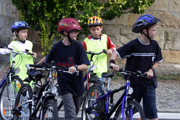 Vier Kinder führen ihre Fahrräder auf einem Bürgersteig entlang einer Steinmauer. Sie tragen Fahrradhelme, zwei Jungen tragen dunkle T-Shirts, während die anderen zwei leuchtende Sicherheitswesten anhaben. Die Szene betont die Wichtigkeit von Sicherheitsausrüstung und achtsames Verhalten im Straßenverkehr, relevant für Unfallprävention und Arbeitsschutzmaßnahmen der Unfallkasse NRW.