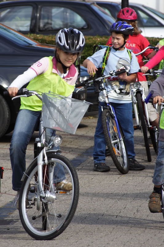 Eine Gruppe von Kindern fährt auf Fahrrädern. Ein Kind im Vordergrund trägt eine helle Schutzweste und einen Helm, während es um eine Kurve fährt. Weitere Kinder im Hintergrund tragen ebenfalls Helme, einige pausieren. Die Szene spielt auf einem Parkplatz mit geparkten Autos im Hintergrund. Das Bild steht im Kontext von Arbeitsschutz und Prävention, gefördert durch die Unfallkasse NRW, um die Bedeutung von Sicherheitsmaßnahmen beim Radfahren zu betonen.
