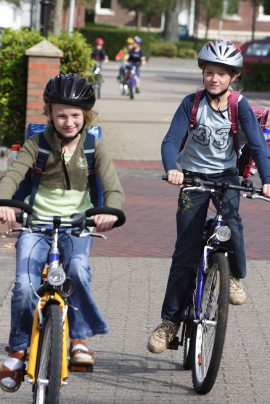 Zwei Kinder, ein Junge und ein Mädchen, fahren mit Helmen auf Fahrrädern eine ruhige Wohnstraße entlang. Sie tragen Rucksäcke und fahren nebeneinander. Weitere Kinder sind im Hintergrund zu sehen. Das Bild vermittelt die Bedeutung von Sicherheit im Straßenverkehr und präventivem Verhalten für die Unfallverhütung, im Kontext des Arbeitsschutzes und der Prävention von Unfällen, relevant für die Unfallkasse NRW.