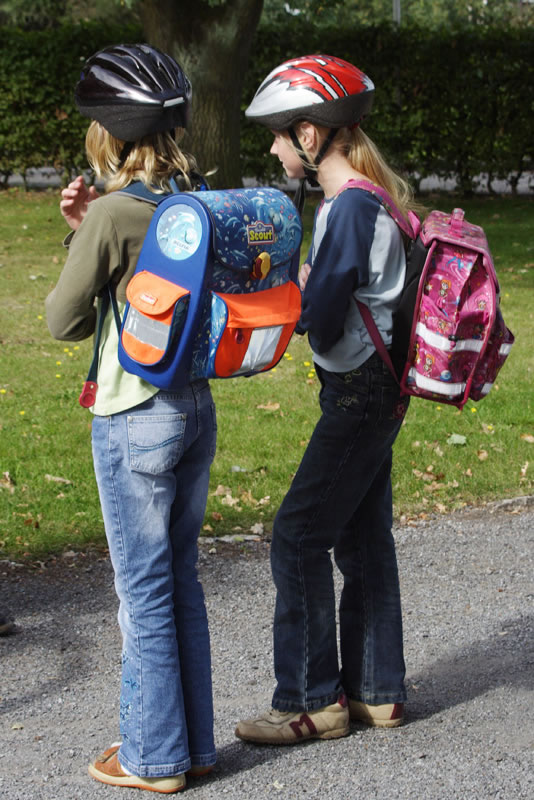 Zwei Kinder auf einem Gehweg, die Fahrradhelme und Schulrucksäcke tragen. Beide tragen Jeans und stehen nebeneinander, nebeneinander. Sie sind in einem parkähnlichen Umfeld mit Gras und Sträuchern im Hintergrund. Dies steht im Kontext von Arbeitsschutz und Prävention, indem die Bedeutung von Sicherheitsmaßnahmen wie dem Tragen von Helmen im Straßenverkehr hervorgehoben wird. Relevanz für Unfallkasse NRW im Rahmen der Sicherheitserziehung.