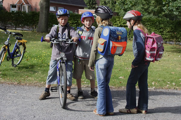 Vier Kinder stehen auf einem gepflasterten Weg auf einem Schulhof. Zwei Jungen, einer mit einem Fahrrad, haben Sicherheitshelme auf und tragen Freizeitkleidung. Zwei Mädchen, ebenfalls mit Helmen, tragen bunte Schultaschen. Im Hintergrund sind Rasenfläche, Bäume und ein Gebäude sichtbar. Das Bild betont die Bedeutung von Sicherheit und Prävention im Schulwegverkehr, in Verbindung mit Unfallkasse NRW.
