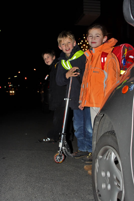 Drei Kinder stehen nachts mit einem Roller auf dem Gehweg neben einem Auto.