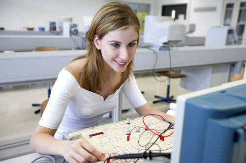Eine junge Frau sitzt in einem modernen Labor und arbeitet an einem elektronischen Schaltkreis. Sie trägt ein weißes Oberteil und schaut auf den Schaltkreis vor ihr, der mit Drähten und elektronischen Bauteilen versehen ist. Im Hintergrund sind verwaschene Laborgeräte und Möbel zu sehen. Diese Szene spiegelt einen sicheren und gut ausgestatteten Arbeitsplatz wider, der den Fokus auf Arbeitsschutz und Prävention legt, was für die Unfallkasse NRW von Bedeutung ist.
