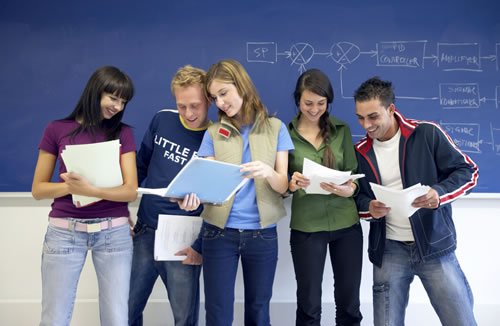 Fünf junge Erwachsene stehen vor einer blauen Tafel und betrachten gemeinsam verschiedene Dokumente. Die Tafel im Hintergrund zeigt schematische Diagramme. Dieses Bild könnte im Kontext von Arbeitsschutz und Präventionsmaßnahmen der Unfallkasse NRW stehen, wobei es das gemeinsame Lernen und Erarbeiten von Sicherheitskonzepten oder Arbeitsrichtlinien symbolisiert.