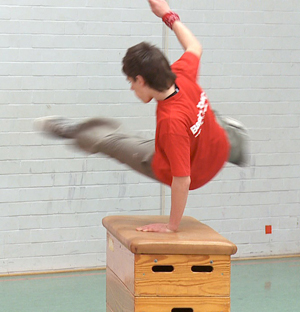 Ein Junge in einem roten T-Shirt führt einen Sprung über einen Turnkasten in einer Sporthalle durch. Er stützt sich mit einer Hand auf den Kasten, während seine Beine in der Luft gespreizt sind. Die Wand im Hintergrund ist hellgrau. Dieses Bild kann im Kontext der Unfallkasse NRW als Hinweis auf die Wichtigkeit von Sicherheit und Prävention im Schulsport interpretiert werden.