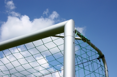 Eine Nahaufnahme der oberen Ecke eines Fußballtors. Das Netz ist grün und fest mit dem Tor verbunden. Im Hintergrund ist ein klarer, blauer Himmel mit vereinzelten weißen Wolken zu sehen. Kontext: Die sichere und stabile Konstruktion von Toren ist wichtig für den Arbeitsschutz und zur Unfallprävention, insbesondere auf Sportplätzen.