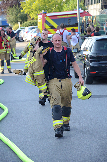 Mehrere Feuerwehrleute, einige in voller Ausrüstung, räumen nach einem Einsatz Schläuche und Ausrüstung auf einer Straße auf. Im Hintergrund steht ein Feuerwehrfahrzeug. Der Fokus liegt auf einem Feuerwehrmann, der Schutzausrüstung und einen Helm trägt. Die Szene vermittelt Arbeitsschutz und Prävention im Rahmen eines Feuerwehreinsatzes für die Unfallkasse NRW.
