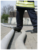 Eine Person in Feuerwehruniform, erkennbar an reflektierenden Streifen, überquert zwei Feuerwehrschläuche, die auf einer gepflasterten Fläche liegen. Im Hintergrund sind unscharf eine Hecke und Gebäude zu erkennen. Die Szene könnte Teil einer Übung oder Sicherheitsdemonstration sein, die auf die Bedeutung von Vorsicht und sicherem Manövrieren in Einsatzsituationen hinweist.