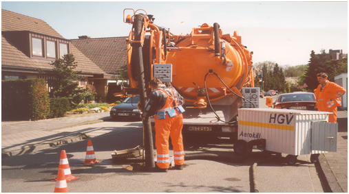 Zwei Arbeiter in orangefarbener Schutzkleidung sichern und bedienen einen großen orangefarbenen Saug-Spülwagen auf einer Wohnstraße. Verkehrsleitkegel sichern die Arbeitsstelle ab. Ein weiterer Arbeiter steht in der Nähe und beobachtet die Szene. Die Veranstaltung wirkt gut organisiert und zeigt die Bedeutung von Sicherheit und Prävention am Arbeitsplatz, was im Kontext der Unfallkasse NRW relevant ist.