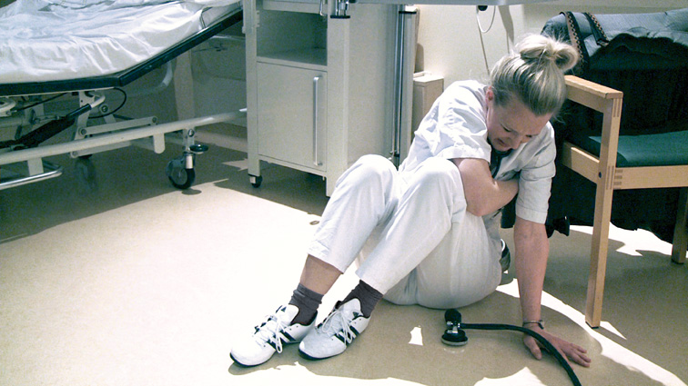 Eine Krankenschwester sitzt verletzt auf dem Boden eines Klinikzimmers. Sie hält ihren Arm und scheint Schmerzen zu haben. Im Raum befinden sich ein Krankenhausbett und medizinisches Equipment. Die Szenerie könnte auf einen Arbeitsunfall hindeuten, was auf die Bedeutung von Arbeitsschutz und Prävention hinweist.