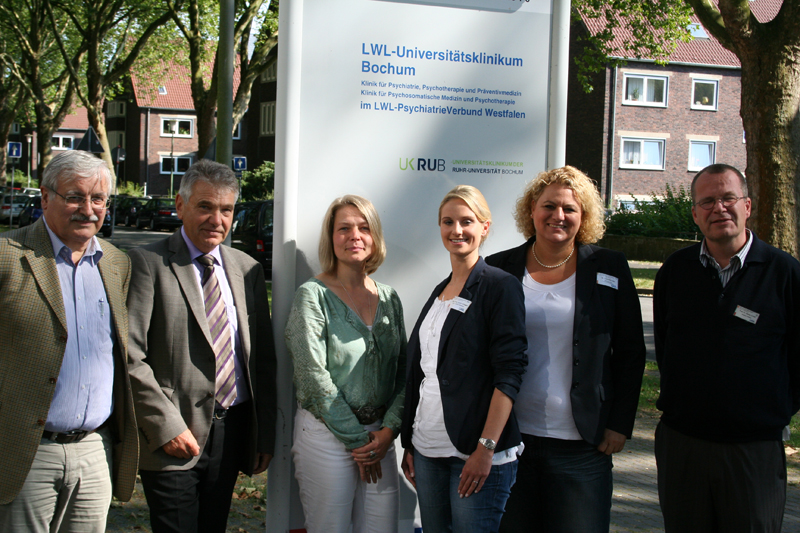 Sechs Personen stehen draußen vor einem Schild der LWL-Universitätsklinik Bochum in einer Baumallee. Das Schild listet verschiedene klinische Abteilungen auf. Die Personen tragen Namensschilder und sind in alltäglicher Kleidung gekleidet. Im Hintergrund sind Wohnhäuser und eine Straße zu sehen. Die Szene spielt bei sonnigem Wetter. Dies könnte im Kontext der Unfallkasse NRW auf Arbeitsschutz und Prävention hinweisen, beispielsweise für ein Treffen oder eine Veranstaltung zur Förderung von Sicherheit am Arbeitsplatz.