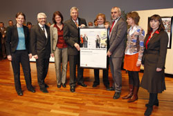 Eine Gruppe von acht Personen, die förmlich gekleidet sind, steht in einem Raum mit Holzboden. Sie halten gemeinsam ein großes Plakat hoch, das vermutlich Informationen über eine Auszeichnung oder ein Projekt im Bereich Arbeitsschutz und Prävention der Unfallkasse NRW enthält. Die Veranstaltung wirkt offiziell, und die Teilnehmer schauen lächelnd oder interessiert in Richtung der Kamera. Im Hintergrund sind Ausstellungstafeln zu sehen.