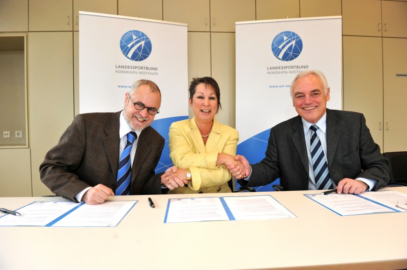 Drei Personen in formeller Kleidung sitzen an einem Tisch und unterzeichnen Dokumente. Zwei Männer und eine Frau mittleren Alters schütteln sich die Hände und lächeln. Im Hintergrund sind Banner des Landessportbundes Nordrhein-Westfalen zu sehen. Die Szene vermittelt eine Partnerschaft im Bereich Arbeitsschutz und Prävention, möglicherweise im Kontext der Unfallkasse NRW.