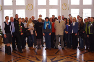 Eine große Gruppe von Männern und Frauen steht in einem Raum mit geometrischem Holzbodenmuster und weißen Wänden mit Fenstern. An der Wand hängen Uhren, die verschiedene Zeiten anzeigen. Einige Personen tragen Namensschilder. Das Bild scheint bei einer Versammlung oder Veranstaltung aufgenommen worden zu sein, möglicherweise im Kontext der Unfallkasse NRW, die sich mit Arbeitsschutz und Prävention befasst.