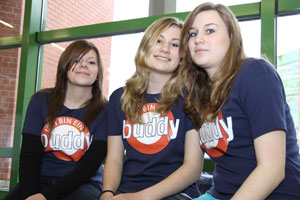 Drei junge Personen sitzen auf einer Bank vor einem Fenster. Sie tragen blaue T-Shirts mit der Aufschrift 'Ich bin ein Buddy'. Die Szenerie wirkt freundlich und einladend, mit Fokus auf Gemeinschaft und Unterstützung. Der Raum ist hell, mit einem roten Backsteinmuster im Hintergrund. Bezug zu einem präventiven Programm zum sozialen Miteinander.