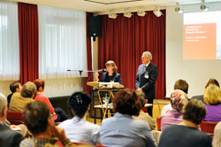 Ein Mann und eine Frau präsentieren an einem Rednerpult vor einem sitzenden Publikum. Der Raum ist mit roten Vorhängen dekoriert, die Präsentation wird auf einer Leinwand rechts gezeigt. Die Personen tragen formelle Kleidung. Der Kontext deutet auf ein Seminar oder eine Schulung zum Thema Arbeitsschutz und Prävention, organisiert von der Unfallkasse NRW, hin. Es sind etwa ein Dutzend Menschen anwesend, die aufmerksam zuhören.