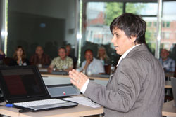 Eine Person in einem grauen Anzug führt eine Präsentation vor einer Gruppe von Menschen in einem Seminarraum durch. Der Raum hat große Fenster, durch die Tageslicht einfällt. Die Zuhörer sitzen aufmerksam und scheinen Notizen zu machen. Im Vordergrund sind ein Laptop und Dokumente auf einem Tisch zu sehen. Dieses Setting könnte eine Schulung oder ein Seminar zur Prävention von Arbeitsunfällen und Arbeitsschutzmaßnahmen im Kontext der Unfallkasse NRW darstellen.