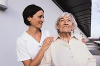 Eine jüngere Frau in weißer Berufskleidung steht lächelnd neben einer älteren Frau. Sie legt die Hand freundlich auf die Schulter der älteren Dame. Die Szene findet im Freien vor einem Gebäude statt. Die Interaktion symbolisiert Fürsorge und Unterstützung, was im Kontext des Arbeitsschutzes und der Prävention relevant für die Unfallkasse NRW ist.