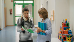 Zwei Frauen stehen in einem hellen Büroflur und scheinen sich zu beraten. Die linke Frau hält ein aufgeschlagenes Dokument, während die rechte Frau ein blau-gestreiftes Notizbuch trägt. Im Hintergrund ist ein buntes, kunstvolles Objekt sichtbar. Ein Brandschutzgerät hängt an der wand. Der Kontext könnte auf Arbeitsschutz oder Präventionsmaßnahmen hinweisen.