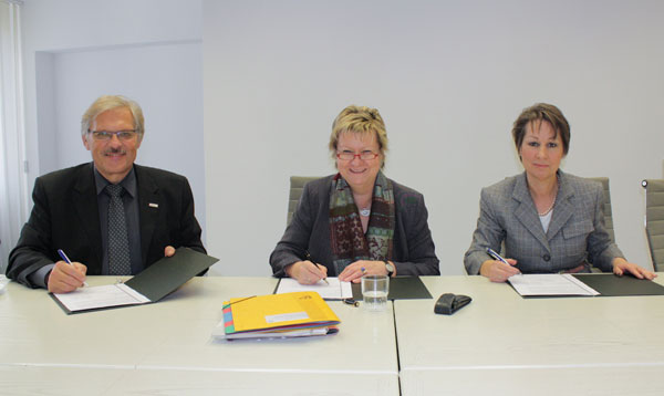 Drei Personen sitzen an einem Tisch und unterzeichnen Dokumente.