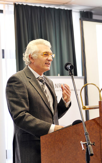 Ein Mann mittleren Alters steht hinter einem braunen Pult und hält eine Rede. Er trägt einen grauen Anzug mit Krawatte und gelbe Brille. Im Hintergrund ist ein Banner der Unfallkasse NRW sichtbar sowie ein Vorhang. Links im Bild ist ein Mikrofon am Pult angebracht. Die Szene vermittelt ein professionelles Umfeld im Kontext von Arbeitsschutz und Prävention.