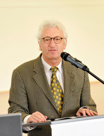 Ein Mann mit grauem Haar und Brille steht an einem Rednerpult und spricht in ein Mikrofon. Er trägt einen karierten Anzug und eine gemusterte Krawatte. Im Vordergrund ist ein Laptop sichtbar. Der Kontext bezieht sich wahrscheinlich auf eine Präsentation zum Thema Arbeitsschutz und Prävention für die Unfallkasse NRW.