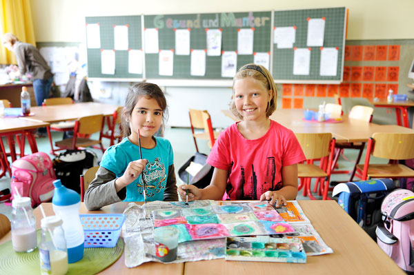 Zwei junge Mädchen sitzen an einem Tisch in einem Klassenzimmer und malen mit Wasserfarben. Sie lächeln und tragen lässige Kleidung. Der Raum ist mit Tischen und Stühlen ausgestattet, im Hintergrund hängen Zeichnungen und Notizen an einer Tafel. Die Szene vermittelt eine freundliche und kreative Lernumgebung, die im Kontext der Unfallkasse NRW auf Sicherheitsaspekte und Prävention im Schulalltag hinweist.