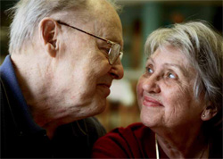 Ein älteres Paar, bestehend aus einem Mann und einer Frau, schaut sich lächelnd und liebevoll in die Augen. Sie sitzen nah beieinander in einem gut beleuchteten Raum. Der Mann trägt eine Brille und ein dunkelblaues Hemd, während die Frau ein dunkelrotes Oberteil trägt. Das Bild vermittelt eine harmonische und liebevolle Stimmung, die den Wert von Beziehungen und sozialem Miteinander unterstreicht.