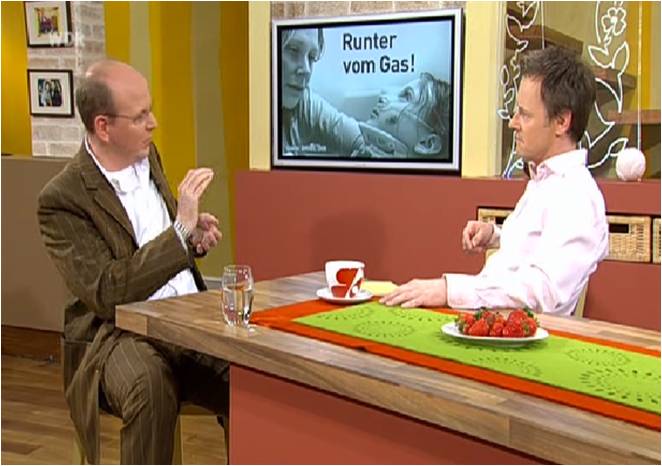 Zwei Männer sitzen in einem Fernsehstudio und führen ein Gespräch. Der Mann links trägt einen braunen Anzug und gestikuliert mit der Hand. Der Mann rechts trägt ein weißes Hemd und hört zu, während er auf einem Holzstuhl sitzt, neben ihm ein Teller mit Erdbeeren und eine Tasse auf einem Tisch. Im Hintergrund ist ein Bildschirm mit der Aufschrift 'Runter vom Gas!' und Bildern von Personen zu sehen. Das Thema scheint sich um Verkehrssicherheit und Prävention zu drehen, im Kontext der Unfallkasse NRW und dem Arbeitsschutz.