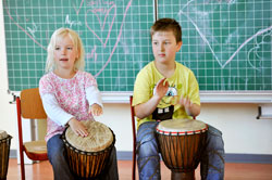 In einem Klassenzimmer sitzen zwei Kinder nebeneinander auf Stühlen und spielen auf Djemben. Sie wirken konzentriert und interessieren sich für die Musikinstrumente. Im Hintergrund ist eine grüne Tafel mit Kreidezeichnungen von Herzen zu sehen. Der Kontext bezieht sich auf den Einsatz von Musik im schulischen Umfeld für präventive und soziale Bildungsmaßnahmen im Sinne der Unfallkasse NRW.