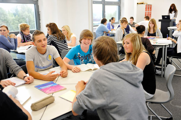 Das Bild zeigt mehrere Jugendliche, die in einem hellen Klassenzimmer in Gruppen an verschiedenen Tischen sitzen und arbeiten. Einige Teilnehmer sind vertieft in ihr Gespräch, während andere Notizen machen. Auf den Tischen liegen Bücher und Schreibmaterialien. Im Hintergrund stehen weitere Schülergruppen und ein Rednerpult. Das Bild vermittelt eine Umgebung des kooperativen Lernens und könnte im Kontext von Arbeitsschutz- und Präventionsmaßnahmen der Unfallkasse NRW als Beispiel dafür dienen, wie wichtig es ist, auch in Bildungseinrichtungen auf eine sichere und förderliche Lernumgebung zu achten.