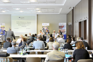Ein Konferenzraum voller Teilnehmer, die einer Präsentation folgen. Im Vordergrund sitzen Menschen an Tischen mit Wasserflaschen. Im Hintergrund ist eine Leinwand mit dem Titel der Veranstaltung, die sich auf Arbeitsschutzmaßnahmen konzentriert. Vor der Leinwand stehen mehrere Redner. Plakate der Unfallkasse NRW sind im Raum verteilt. Das Setting vermittelt eine professionelle Atmosphäre und thematisiert Sicherheitsaspekte und Prävention in der Arbeitswelt.