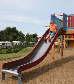 Ein Kind in einem orangefarbenen T-Shirt rutscht lächelnd eine breite, geschwungene Rutsche hinunter. Der Spielplatz ist umgeben von Wohnwagen und Bäumen im Hintergrund. Der Boden besteht aus Holzschnitzeln für eine weiche Landung. Das Bild betont die Wichtigkeit von Sicherheitsmaßnahmen und geeignetem Untergrund bei Spielplätzen, ein Kernthema für die Unfallkasse NRW im Bereich Arbeitsschutz und Prävention.