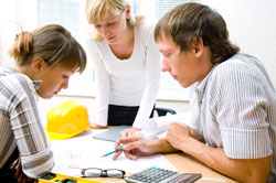 Drei Personen, zwei Männer und eine Frau, besprechen Bauzeichnungen an einem Tisch. Ein gelber Schutzhelm liegt auf dem Tisch, was auf Arbeitssicherheit hinweist. Ein Taschenrechner liegt ebenfalls auf dem Tisch, was auf präzise Planung und Berechnung hindeutet. Die Besprechung könnte im Kontext von Arbeitsschutz und Prävention bei Bauprojekten bei der Unfallkasse NRW stehen.